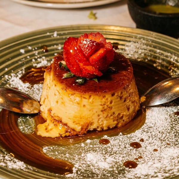 Caramel flan topped with a strawberry rose, powdered sugar, and chocolate sauce on a decorative plate.