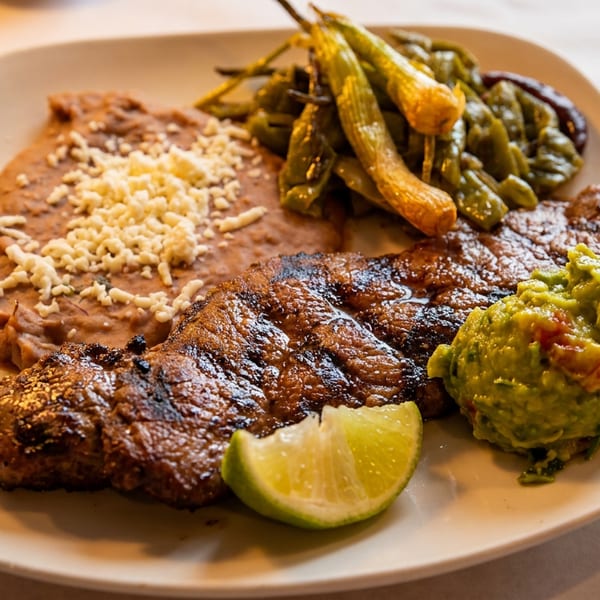 Grilled steak served with refried beans, sautéed green vegetables, guacamole, cheese, and a lime wedge on a plate.