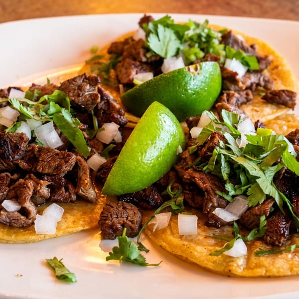 Two crispy beef tacos topped with diced onions and cilantro, garnished with lime wedges on a white plate.
