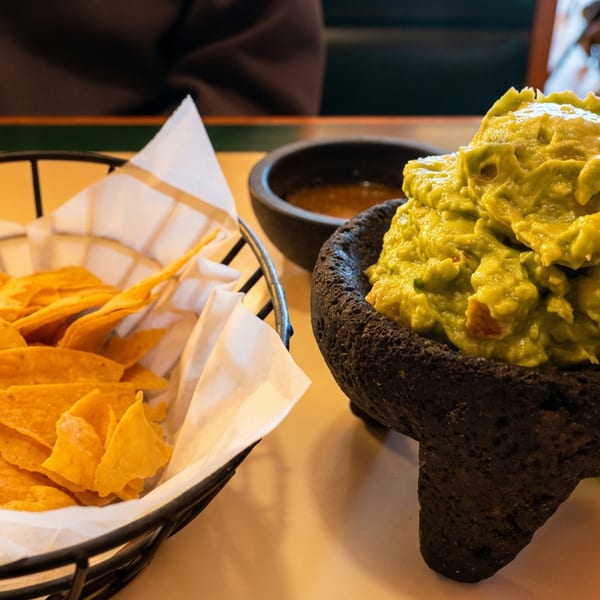 Basket of tortilla chips beside freshly prepared guacamole in a black stone molcajete with dipping sauces at a restaurant table.