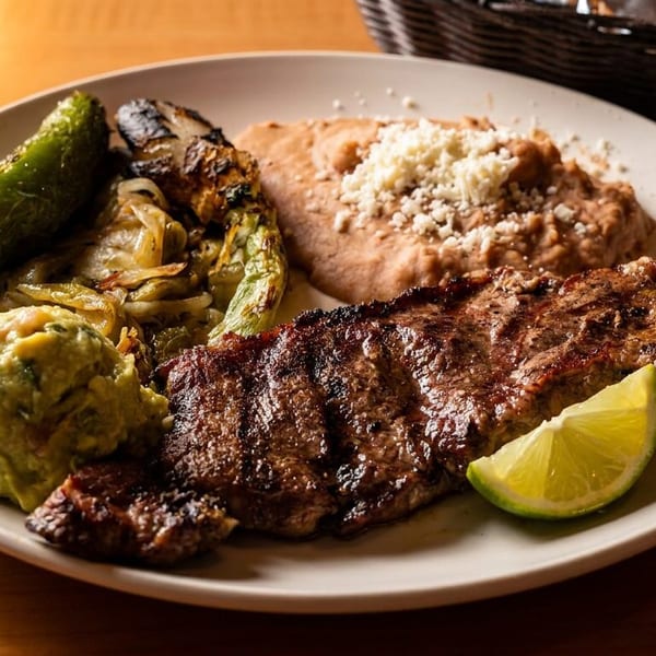 Grilled steak served with guacamole, roasted vegetables, refried beans, and a lime wedge on a plate.